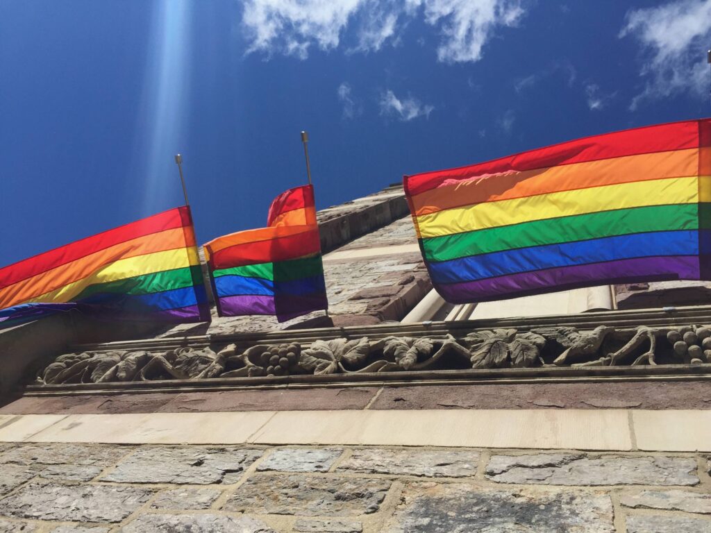 Rainbow pride flags flying from a carved stone building against a blue sky