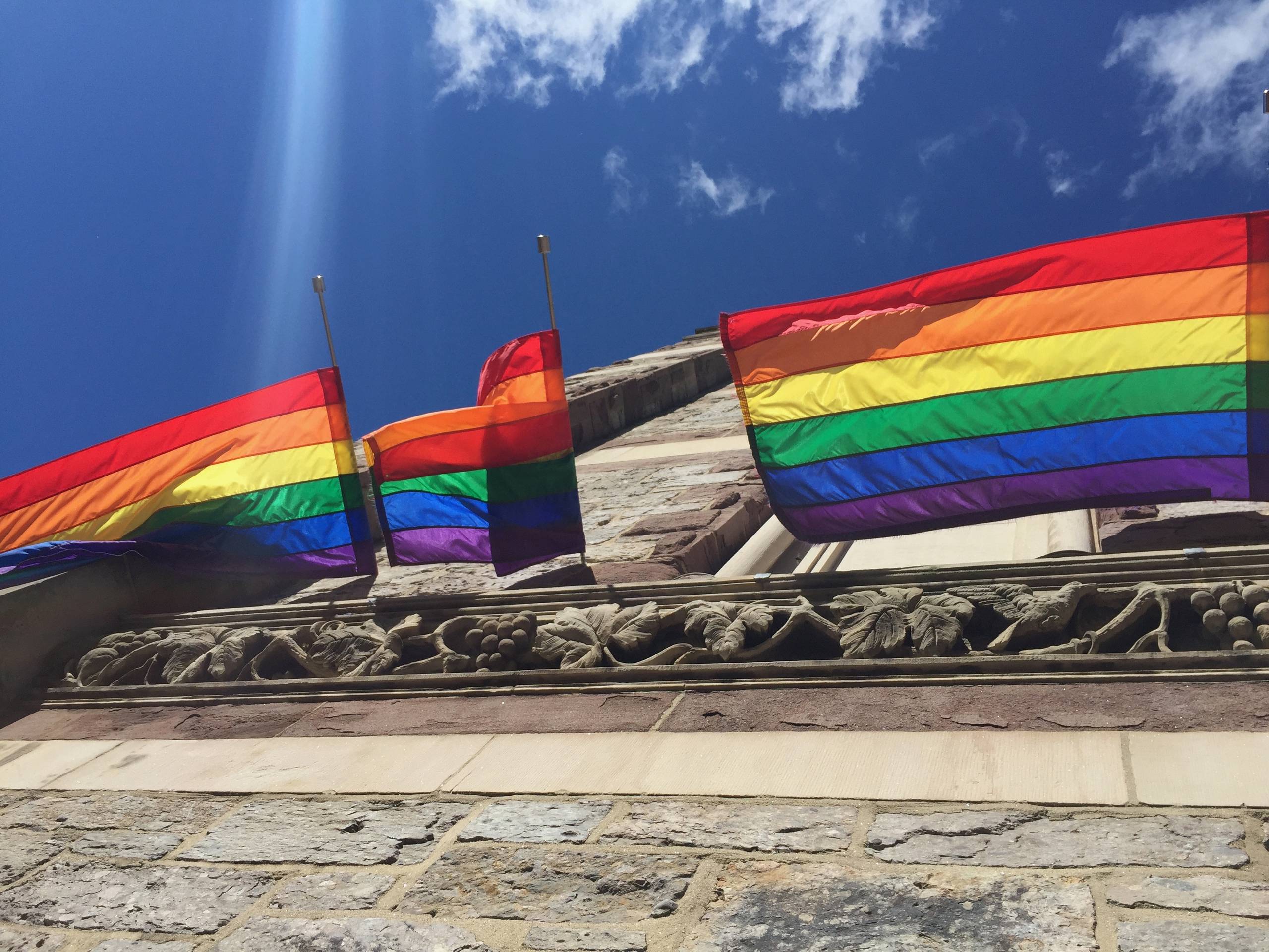 Rainbow pride flags flying from a carved stone building against a blue sky